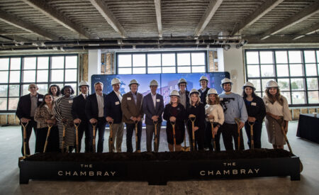 A group of people pose for a photo with hardhats on and shovel in dirt.